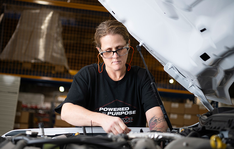A female technician wearing safety equipment performing maintenance on a white vehicle in a workshop environment.
