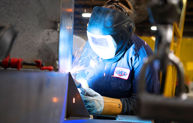 A close-up view of a welder at work in a manufacturing facility, wearing full personal protective equipment.