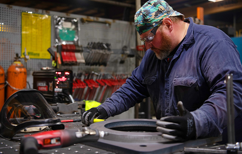 A man wearing safety gear working on a large circular metal component on a workbench in a manufacturing facility.