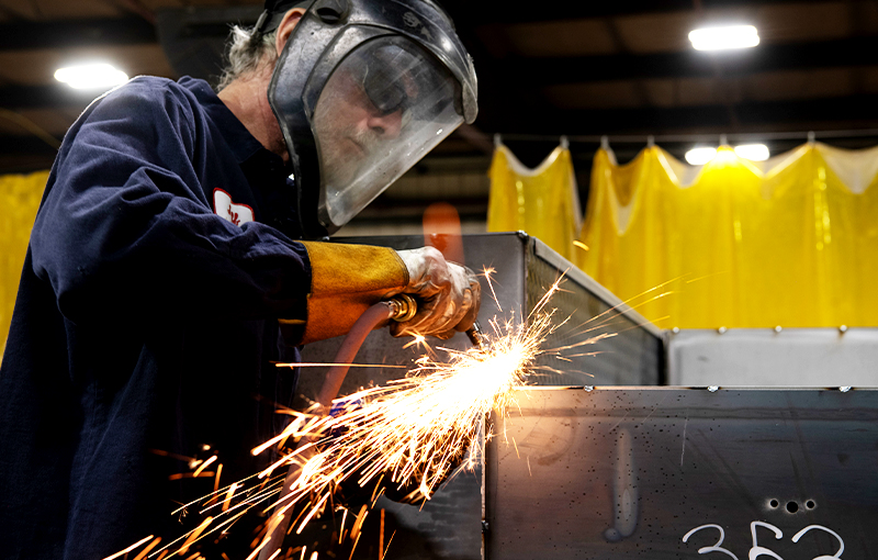A metalworker grinding a steel component with sparks flying with a full-face shield and gloves.