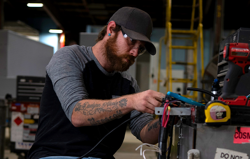 A technician wearing safety glasses works on an electrical wiring harness at a workbench in a manufacturing facility.