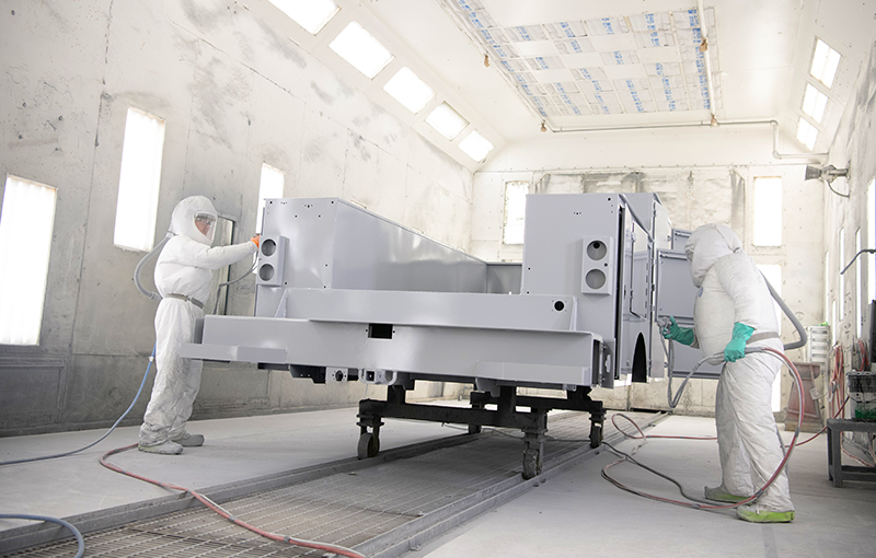 Two technicians wearing full white protective suits and respirators use spray painting gray primer or coating to a large truck body inside a bright industrial spray booth.