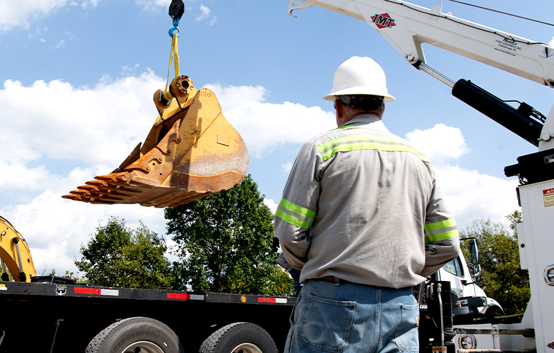 A worker in a white hard hat and high-visibility shirt supervises an IMT service crane as it lifts a large, yellow excavator bucket onto the back of a flatbed truck.