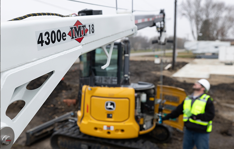 A close-up of an IMT 4300e service crane boom in the foreground, with an operator in a safety vest standing next to a yellow mini-excavator at a job site.
