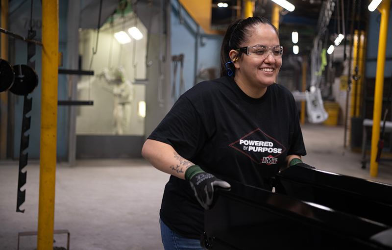 A woman wearing safety equipment smiling while moving black industrial equipment through a manufacturing plant.